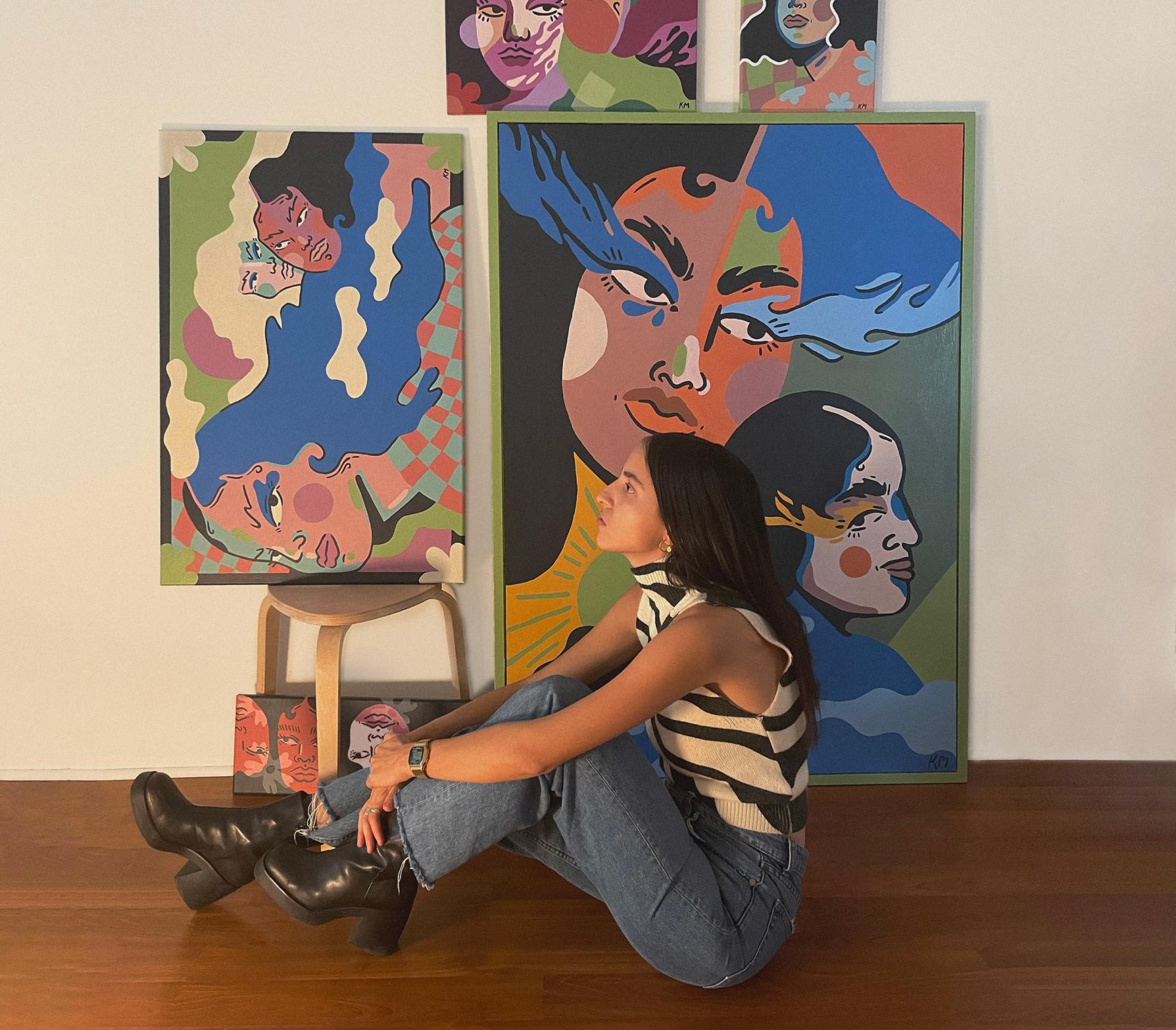 
        Photo of artist Kelly Malka sitting on the floor,
        in front of some of her artwork.
      