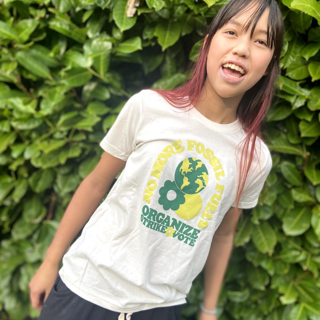A photo of a young girl wearing her organize t-shirt. She’s in front of a hedge and smiling.