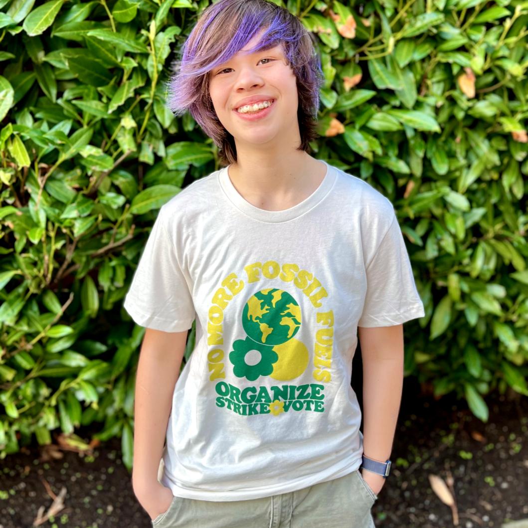 A photo of a young person wearing their organize t-shirt. They’re smiling and standing in front of a hedge.